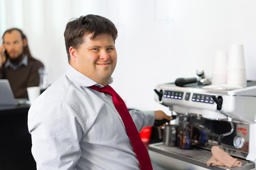 a-happy-man-at-a-coffee-machine-7697916 Man with Down syndrome smiling while making coffee at a cafe, symbolizing inclusivity.